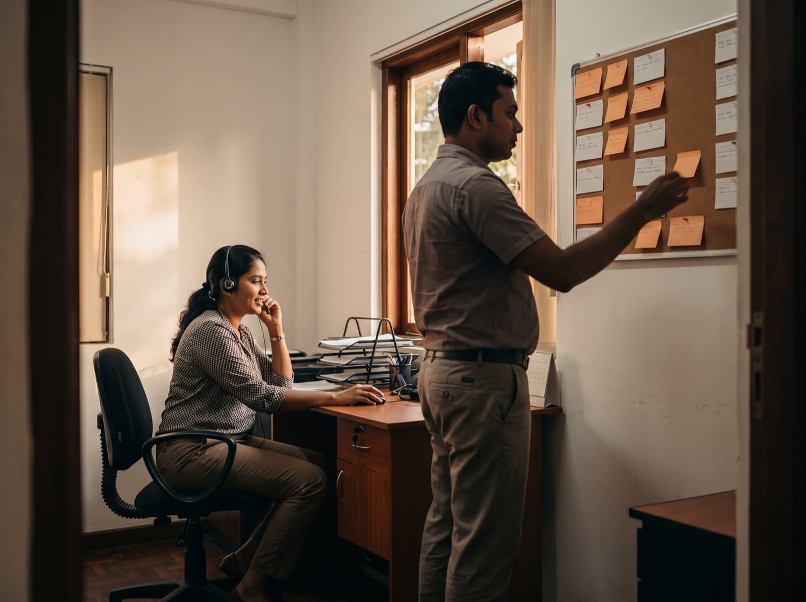 Two colleagues working a pipeline board of sticky notes and a support call.